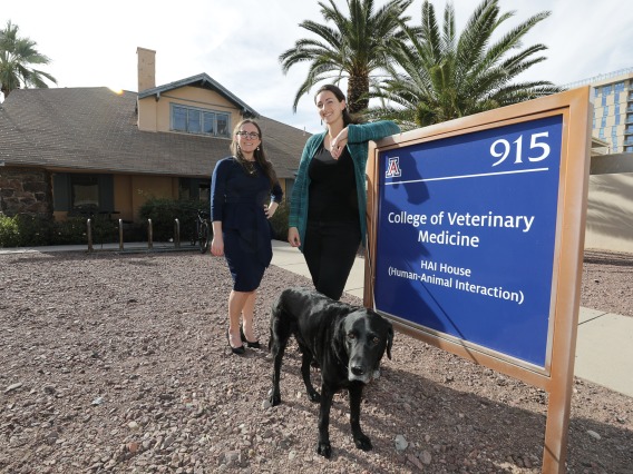 Dr. Maggie O'Haire and Sarah Leighton pose together in front of a sign that reads, "College of Veterinary Medicine Human-Animal Interaction House." The house, a research building, stands in the background.