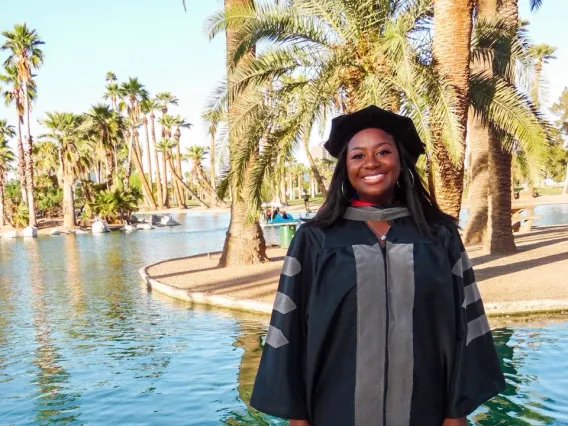 Ari Adams stands in graduation regalia in front of a water feature and palm trees.