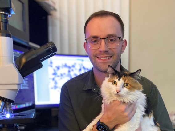 Jeremy Bessett sits in front of a microscope and holds a calico cat.