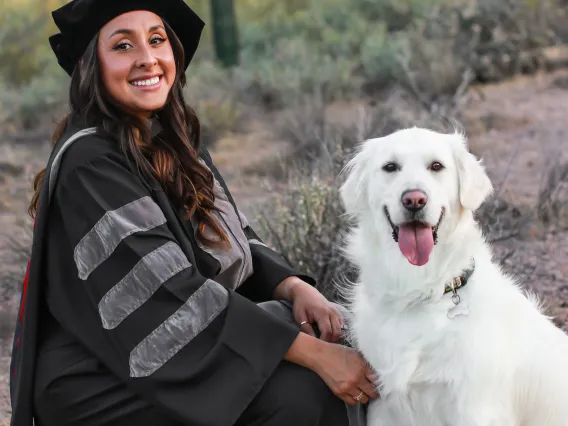 Camila Rocha wears graduation regalia and poses with a light-colored dog.