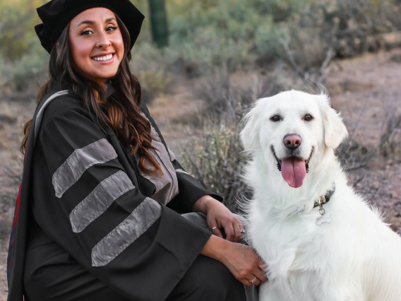 Camila Rocha wears graduation regalia and poses with a light-colored dog.