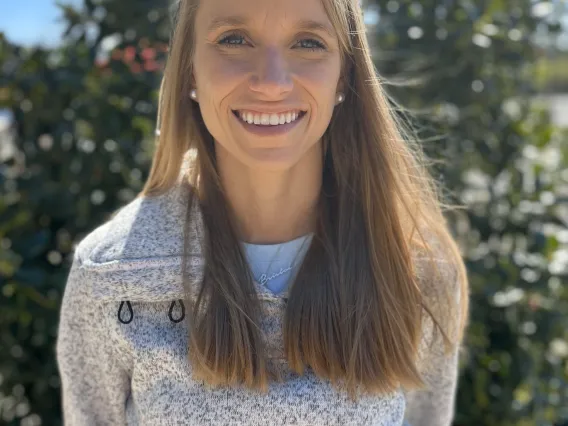 Woman with blond hair and grey sweater standing in front of a tree.