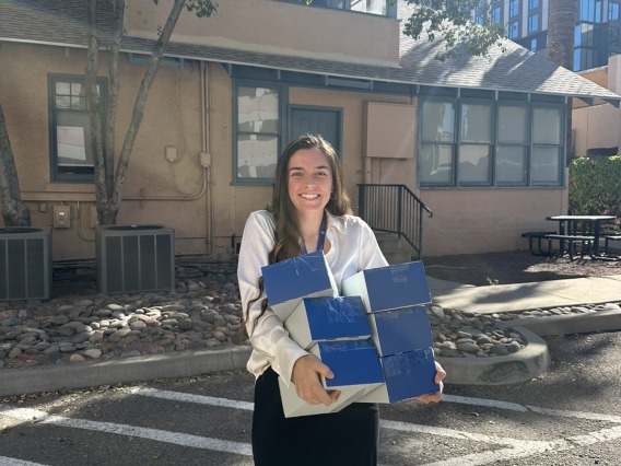 A woman stands in front of the Human-Animal Interaction research house and holds an armload of test kits in blue boxes.