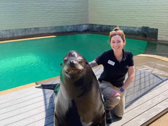 A woman smiles and kneels next to a large seal near a tank of water.