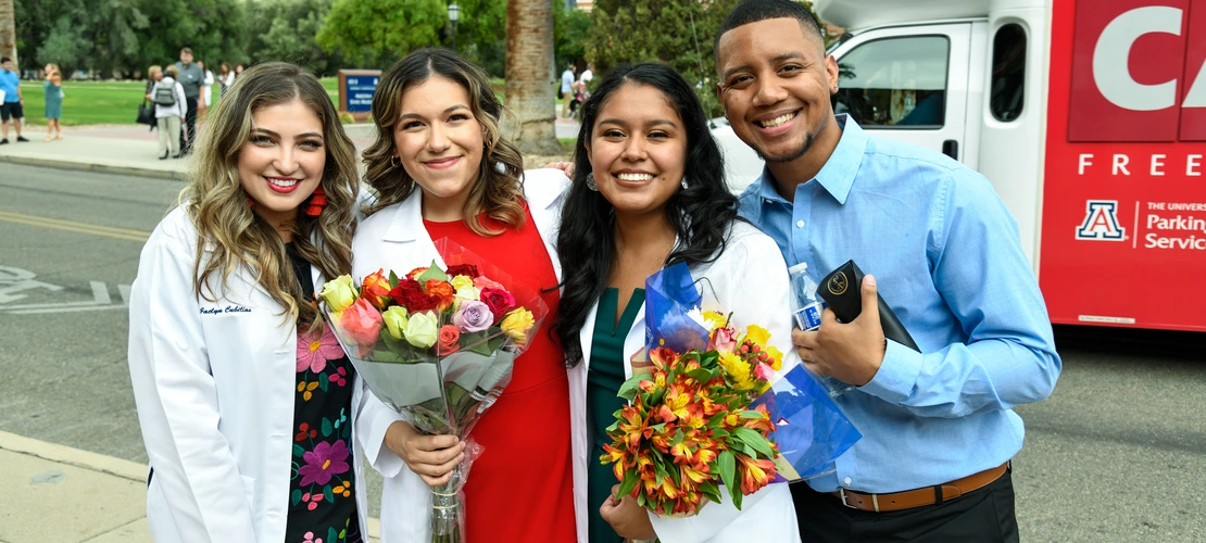 A group of four students in white coats poses outdoors.