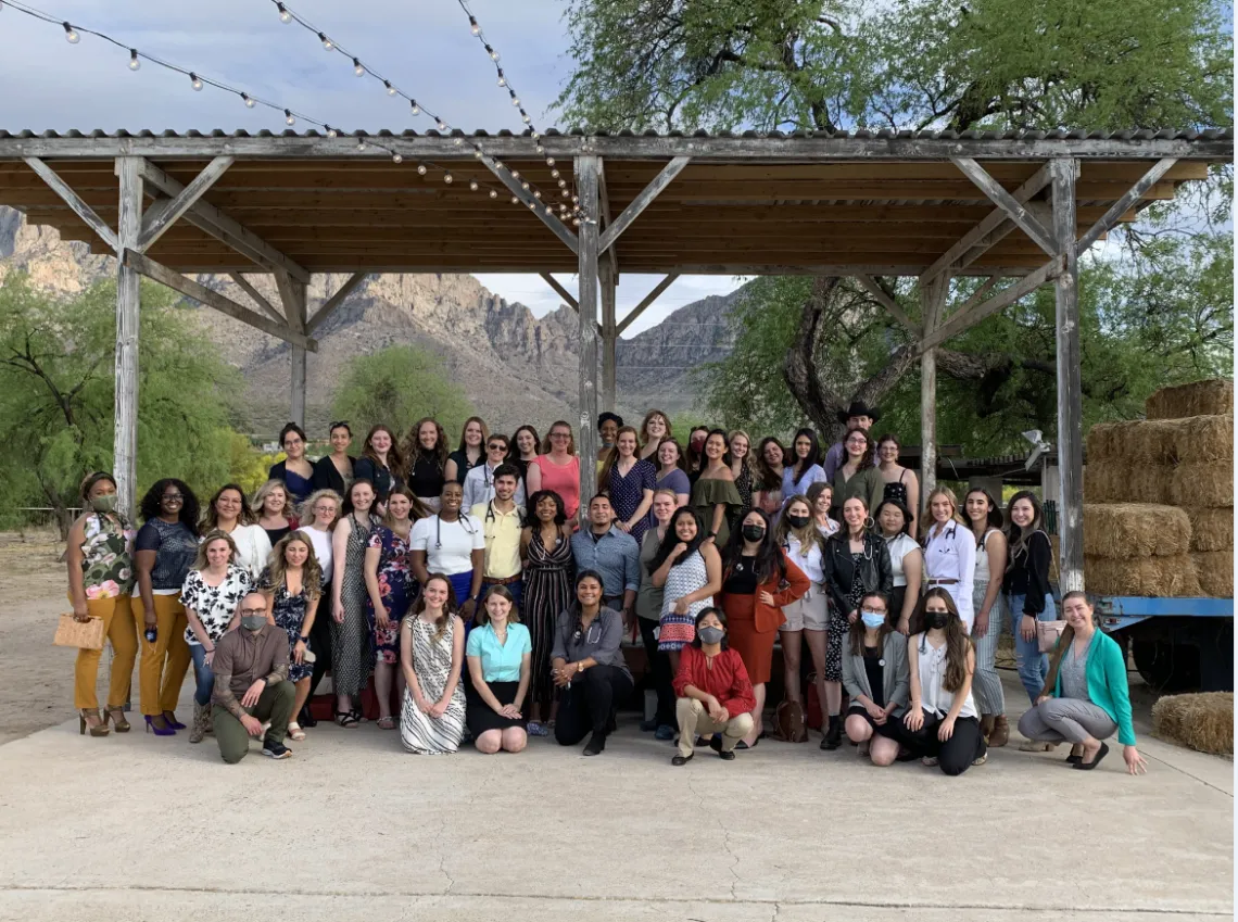 A group of over 50 students stand together. In the background are mountains and to the right side is a stack of hay bales.