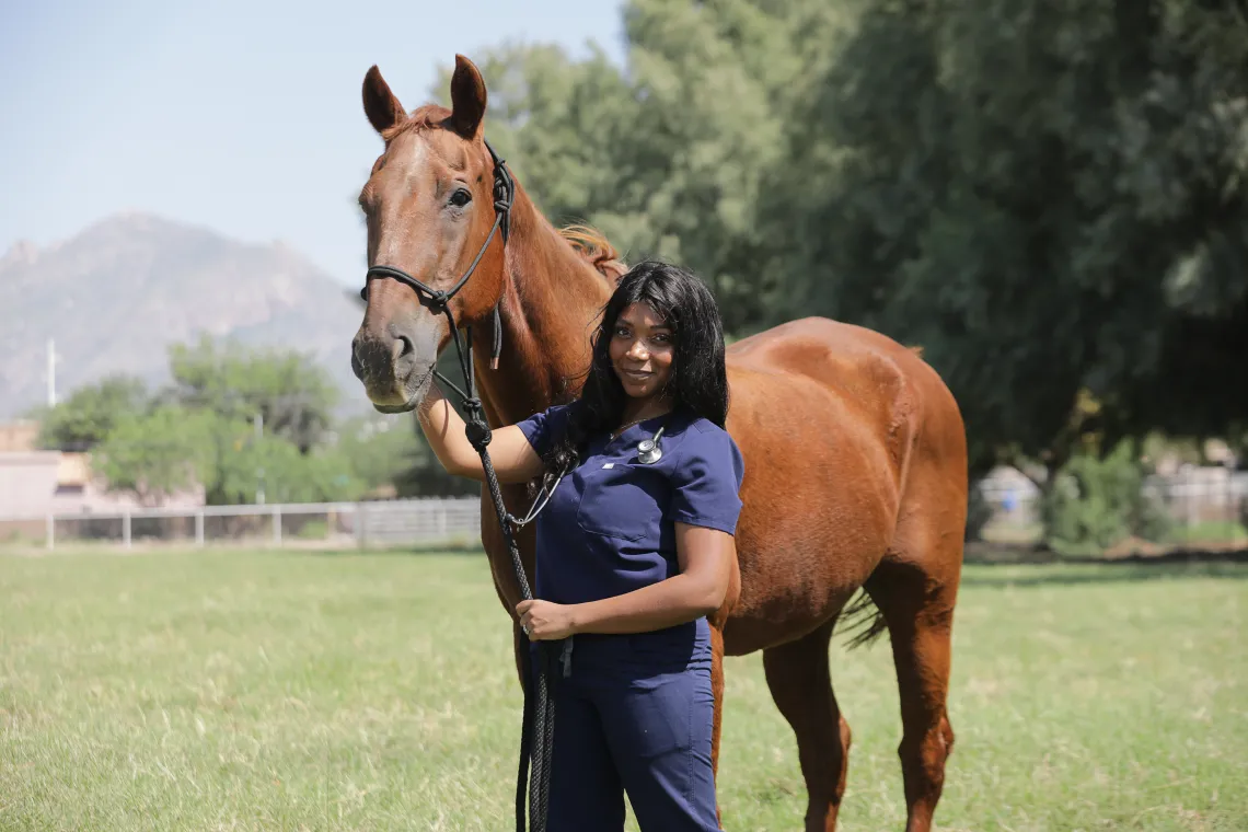 A young woman and a horse look toward the camera. The woman holds a rope in one hand and touches the horse's face with the other.