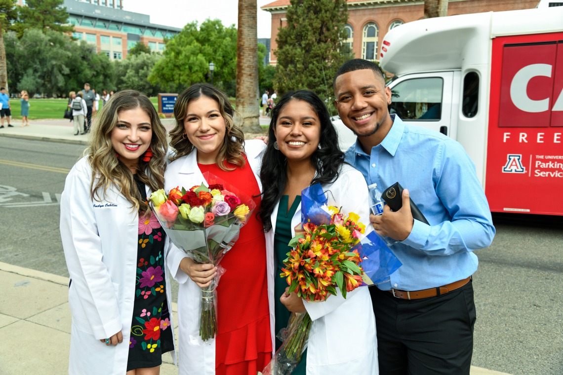 A group of four students in white coats poses outdoors.