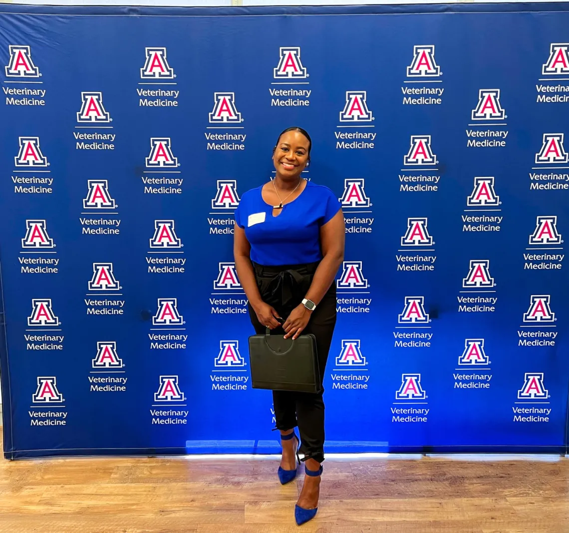 A student poses in front of a wall patterned with the University of Arizona College of Veterinary Medicine logo.