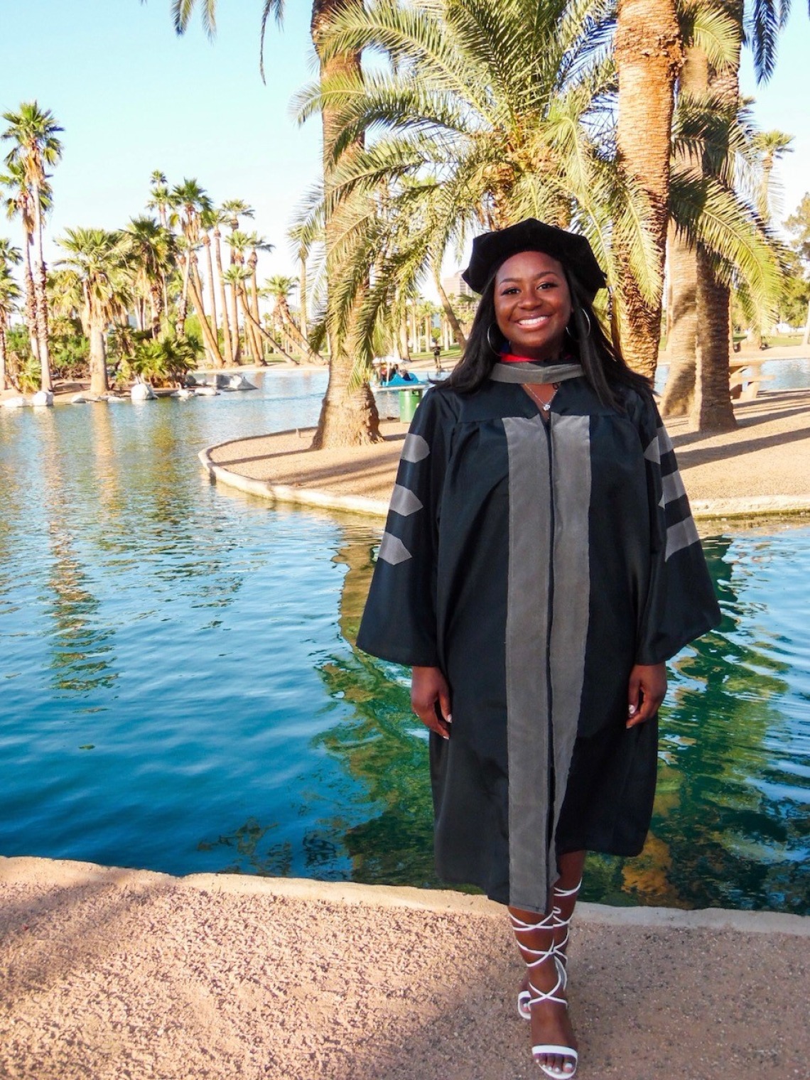Ari Adams stands in graduation regalia in front of a water feature and palm trees.