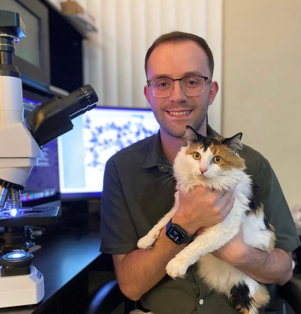 Jeremy Bessett sits in front of a microscope and holds a calico cat.