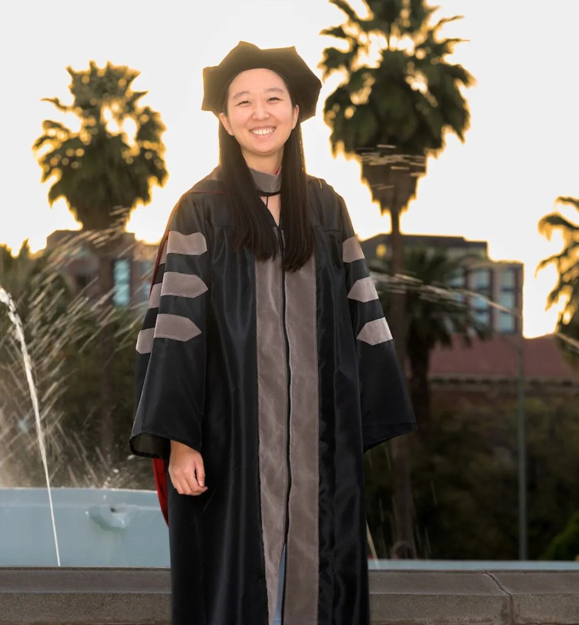 Annika He wears her graduation regalia and stands in front of a fountain and palm trees.