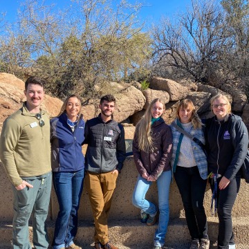 Five veterinary students and one professor stand in front of a bobcat enclosure, in which a bobcat naps on a ledge.
