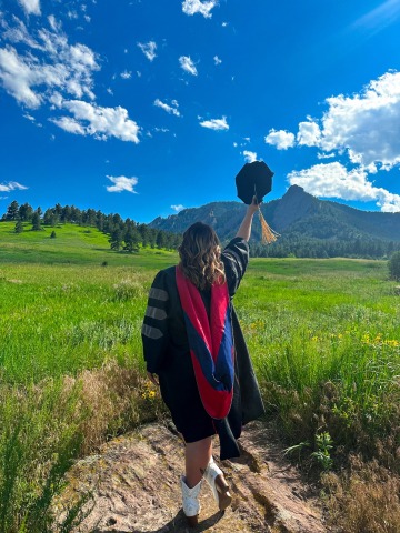 Student Haley McCarthy poses in front of a mountain landscape, wearing graduation regalia. She holds her cap in the air.