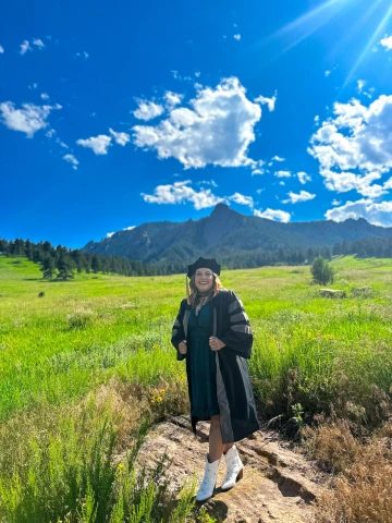 Student Haley McCarthy poses in front of a mountain landscape, wearing graduation regalia.