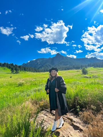 Student Haley McCarthy poses in front of a mountain landscape, wearing graduation regalia.