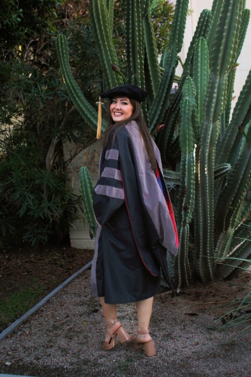 Alexis Tostado wears graduation regalia and poses in front of a large cactus.