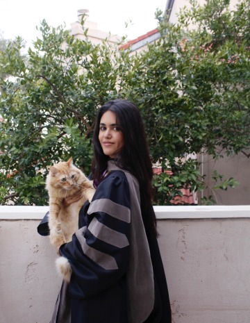 Shannon Khodadad wears graduation regalia and holds an orange cat.