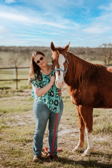 Dr. Megan Owen, wearing sunglasses, a green shirt, and jeans, poses with a brown horse with a white face.