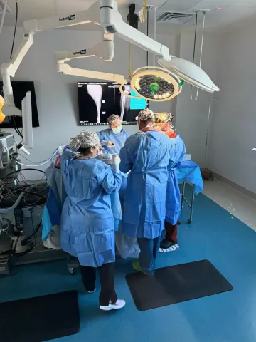 Three individuals in surgical gowns participate in a surgery, with their backs to the camera and x-rays visible on the wall in front of them.