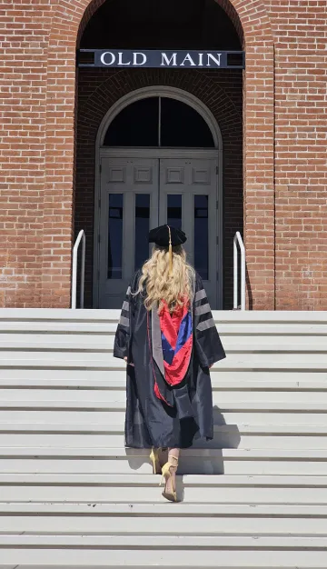 A woman dressed in doctoral regalia walks up the steps to Old Main, with her back to the camera.