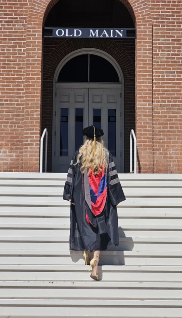 A woman dressed in doctoral regalia walks up the steps to Old Main, with her back to the camera.