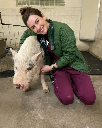 A woman smiles and kneels next to a pig.