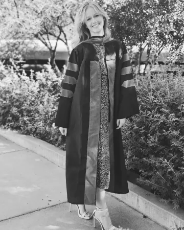 Black and white image of a woman in doctoral regalia standing in front of foliage and smiling.